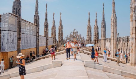 Visitors stroll between spires on the rooftop of Milan Cathedral, some seeking shade under hats or umbrellas during a hot summer day