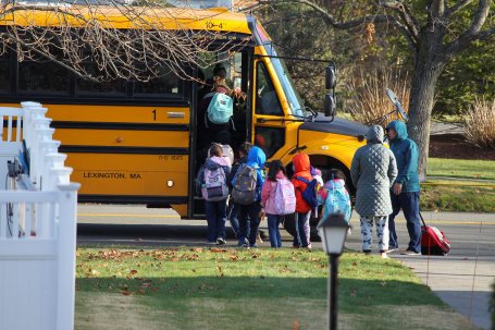 Children wearing backpacks board a yellow school bus while two adults stand nearby.