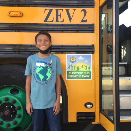 A child smiling in front of a school bus. On the bus, there is a sign that says "Electric Bus. Breathe Easy."
