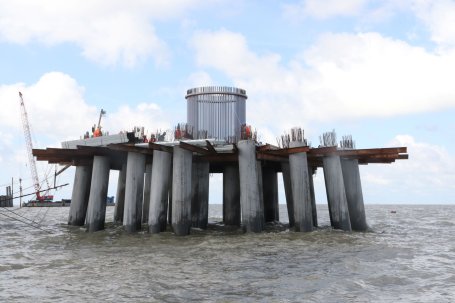 Large cement pillars with a platform holding workers building a wind turbine extend out of the ocean.