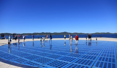 People walk across in-ground solar panels on a seaside plaza on a sunny day.