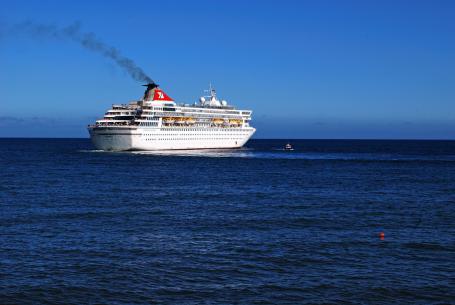 A large cruise ship sailing on bright blue water with dark fumes emitting from its smoke stack.