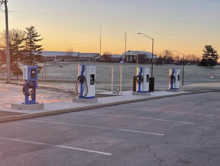 Four electric vehicle charging stations installed in a school parking lot at sunrise, with a school building in the background.