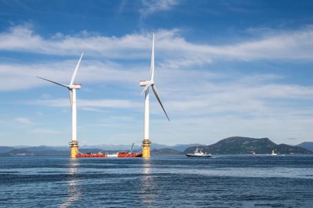 Two massive wind turbines floating offshore with mountains in the background.