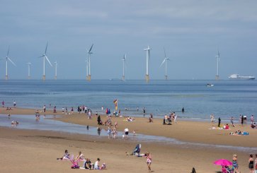 Groups of people are scattered across a large, sandy beach on a sunny day with floating wind turbines in the background.