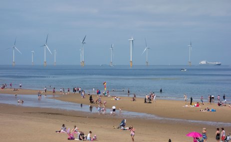 Groups of people are scattered across a large, sandy beach on a sunny day with floating wind turbines in the background.