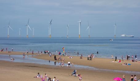 Groups of people are scattered across a large, sandy beach on a sunny day with floating wind turbines in the background.