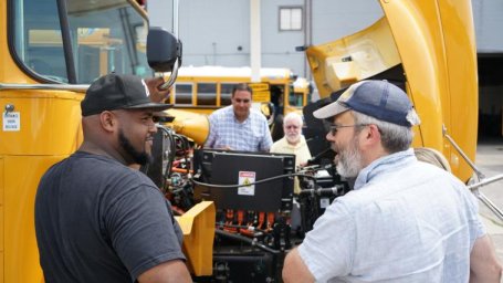 People looking inside the front hood of a yellow electric school bus.