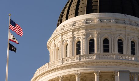 A flagpole with three flags including U.S. and California, stands next to the dome of the California State Capitol building in Sacramento.