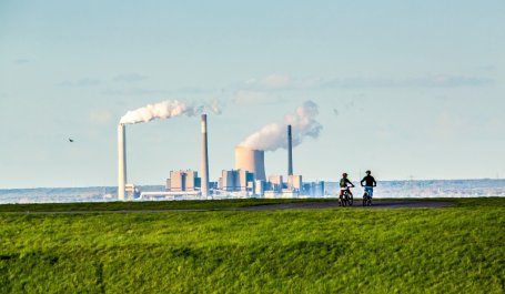 View of two mountain bikers on a grassy hillside with a coal plant in the distance.