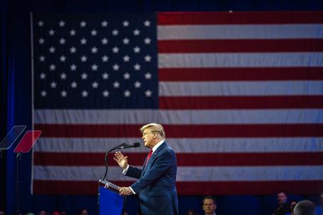 Donald Trump onstage at a podium in front of a large American flag.