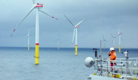 A worker in an orange jumpsuit and white hard hat looks off the side of a ship toward large wind turbines in the ocean.