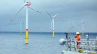 A worker in an orange jumpsuit and white hard hat looks off the side of a ship toward large wind turbines in the ocean.