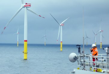 A worker in an orange jumpsuit and white hard hat looks off the side of a ship toward large wind turbines in the ocean.