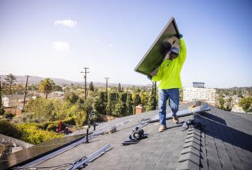 A construction worker carries a solar panel across a house roof for installation